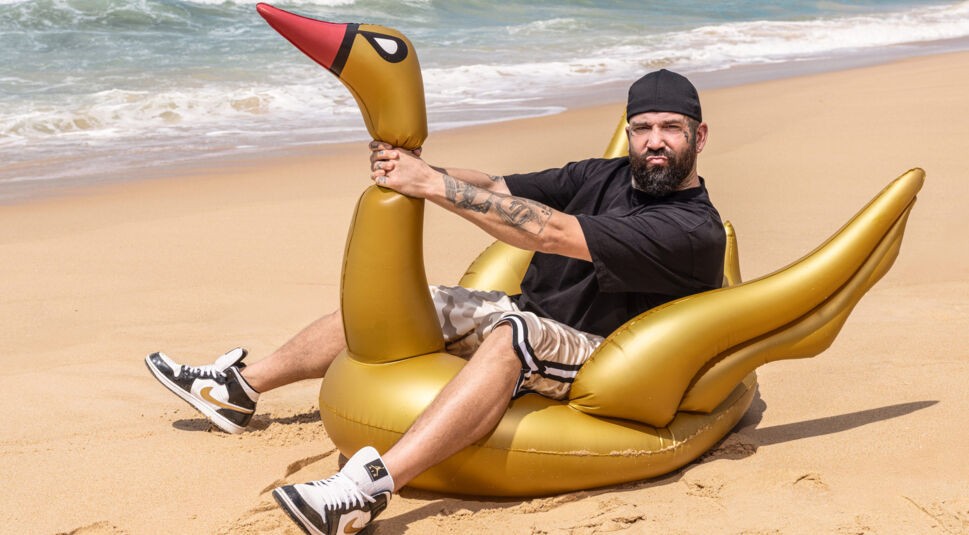 Auf dem Bild zur Reality-Show „Promis unter Palmen 2025“ ist Chris Manazidis zu sehen, wie er auf einem goldenen, aufblasbaren Schwantier am Strand sitzt. Er trägt ein schwarzes Shirt, camouflagemusterte Shorts und schwarz-weiße Sneaker. Mit einem verschmitzten Gesichtsausdruck posiert er lässig vor der Kulisse aus Sand, Meer und blauem Himmel, was der Szene eine humorvolle Note verleiht.