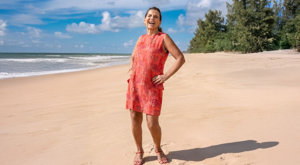 Auf dem Bild zur Reality-Show „Promis unter Palmen 2025“ ist Claudia Obert zu sehen, wie sie lachend am Strand posiert. Sie trägt ein sommerliches, rotes Kleid mit tropischem Muster und rote Sandalen. Im Hintergrund erstreckt sich ein idyllischer Strand mit blauem Himmel, Meereswellen und grünen Bäumen. Ihre entspannte Haltung vermittelt Lebensfreude.