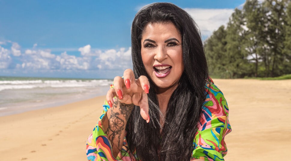 Auf dem Bild zur Reality-Show „Promis unter Palmen 2025“ ist Iris Klein am Strand zu sehen. Sie trägt ein buntes, gemustertes Outfit und macht eine spielerische Geste mit ihren rot lackierten Fingernägeln in die Kamera. Ihre schwarzen Haare fallen offen über die Schultern, und sie lächelt lebhaft. Im Hintergrund sind der Sandstrand, das Meer und ein klarer Himmel zu erkennen.
