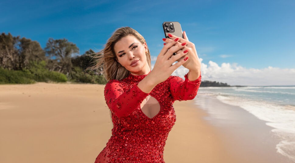 Auf dem Bild zur Reality-Show „Promis unter Palmen 2025“ ist Kim Virginia zu sehen, wie sie am Strand ein Selfie aufnimmt. Sie trägt ein auffälliges, rotes Kleid mit funkelnden Details und hat perfekt gestylte Haare. Ihre rote Maniküre passt zu ihrem glamourösen Look. Im Hintergrund sind Sandstrand, Meer und ein strahlend blauer Himmel zu erkennen, die eine luxuriöse Atmosphäre schaffen.