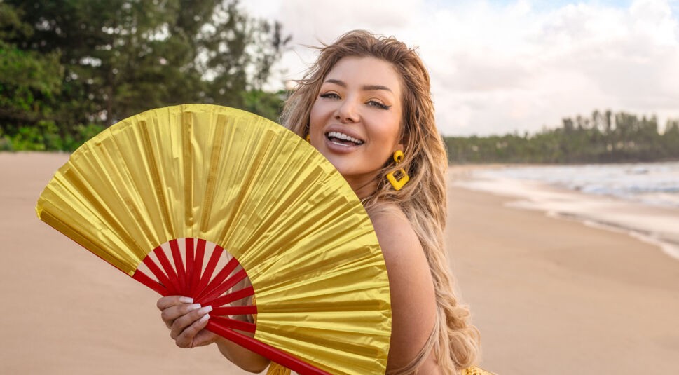 Auf dem Bild zur Reality-Show „Promis unter Palmen 2025“ ist Melody Haase am Strand zu sehen. Sie hält einen gelb-roten Fächer vor sich und strahlt in die Kamera. Ihr sommerliches Outfit und gelbe Ohrringe passen perfekt zur tropischen Kulisse. Im Hintergrund sind der Sandstrand, das Meer und grüne Bäume unter einem hellen Himmel zu erkennen, was eine lebhafte und fröhliche Stimmung vermittelt.