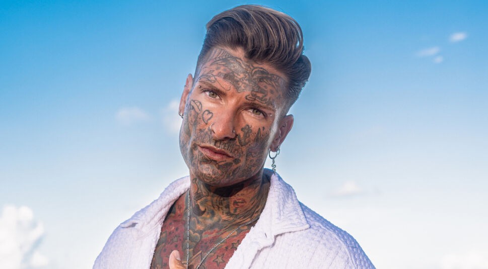 Auf dem Bild zur Reality-Show „Promis unter Palmen 2025“ ist Nikola Glumac zu sehen. Er trägt ein weißes Hemd, das offen seinen tätowierten Oberkörper zeigt. Sein Gesicht ist ebenfalls vollständig tätowiert, was seinen Look einzigartig macht. Mit leicht geneigtem Kopf und ernstem Blick posiert er vor einem klaren blauen Himmel, was die Szene minimalistisch und ausdrucksstark erscheinen lässt.