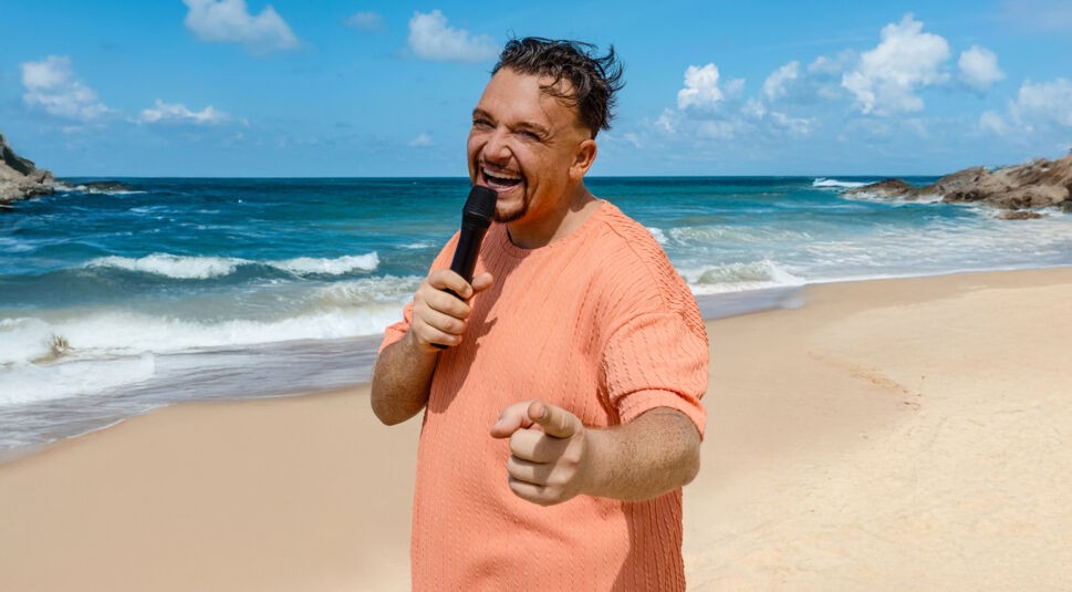 Auf dem Bild zur Reality-Show „Promis unter Palmen 2025“ ist Menowin Fröhlich zu sehen, wie er lächelnd ein Mikrofon hält und in die Kamera zeigt. Er trägt ein lachsfarbenes Oberteil und steht an einem sonnigen Strand mit türkisfarbenem Meer und Wellen im Hintergrund. Seine energiegeladene Pose strahlt gute Laune und Selbstbewusstsein aus.