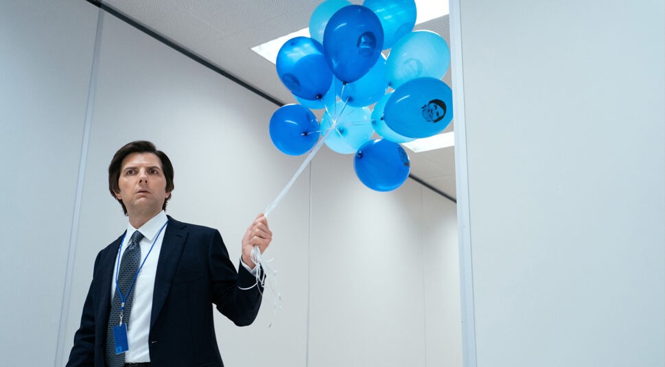 Auf dem Bild zu Severance Staffel 3 ist Mark Scout, gespielt von Adam Scott, zu sehen. Er trägt einen dunklen Anzug mit Krawatte und ein Namensschild um den Hals. In der rechten Hand hält er eine Gruppe blauer Luftballons, von denen einige ein Gesicht aufgedruckt haben. Die Szene spielt in einem sterilen Büroflur mit weißen Wänden und gedecktem Licht, was eine geheimnisvolle und angespannte Atmosphäre vermittelt.