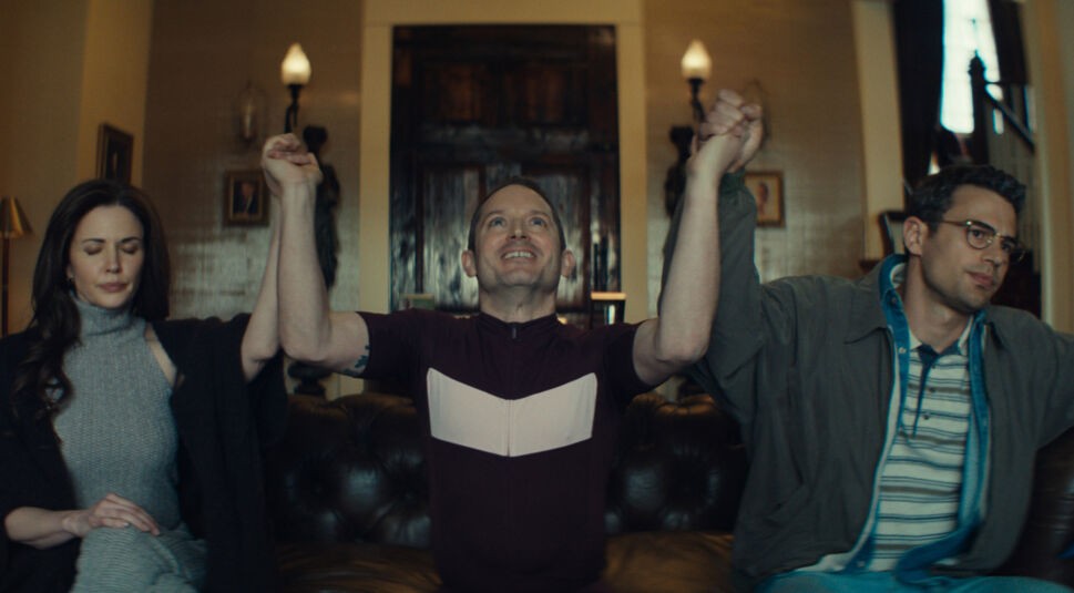 In einer Szene aus "The Monkey" sitzen Laura Mennell, Elijah Wood und Theo James auf einem Ledersofa. Laura Mennell sitzt links mit geschlossenen Augen in einer ruhigen Haltung. In der Mitte hebt Elijah Wood triumphierend die Hände der beiden anderen in die Luft und lächelt. Rechts schaut Theo James mit einem nachdenklichen Ausdruck. Die Szene wirkt symbolisch und emotional intensiv.