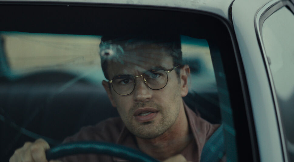 In einer Szene aus "The Monkey" ist Theo James hinter der Windschutzscheibe eines Autos zu sehen. Er trägt eine Brille und schaut konzentriert nach vorne, während er das Lenkrad hält. Eine Einschussstelle in der Scheibe deutet auf eine gefährliche Situation hin. Die Atmosphäre wirkt angespannt und dramatisch.