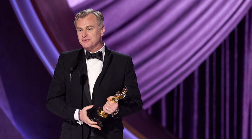 Auf dem Bild in dem Artikel zu „The Odyssey“ ist Christopher Nolan zu sehen, wie er auf einer Bühne in einem eleganten schwarzen Smoking und mit einer Fliege steht. In der rechten Hand hält er eine goldene Trophäe. Der Hintergrund ist mit violetten Vorhängen gestaltet und wirkt feierlich und glamourös.