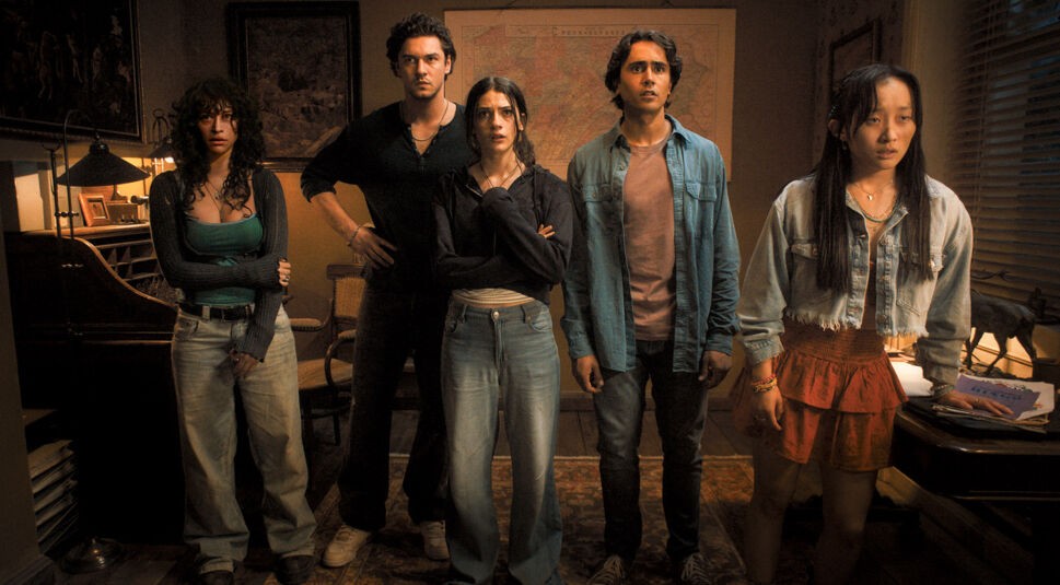 Im Bild zum Until Dawn-Film stehen fünf Charaktere nebeneinander in einem gemütlich eingerichteten Raum mit gedämpfter Beleuchtung. Von links nach rechts sind Nina (Odessa A'zion), Abel (Belmont Cameli), Clover (Ella Rubin), Max (Michael Cimino) und Megan (Ji-young Joo) zu sehen. Alle wirken angespannt und fokussiert, während sie offenbar eine bedrohliche Situation beobachten. Eine Karte im Hintergrund und ein Schreibtisch rechts im Bild verstärken die geheimnisvolle Atmosphäre.