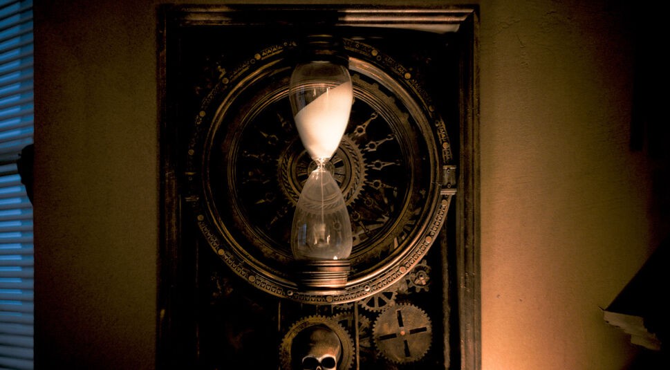 Eine symbolträchtige Sanduhr vor einem mechanischen Hintergrund mit Zahnrädern und einem Totenkopf ist im Bild zum Until Dawn-Film zu sehen. Die Sanduhr, deren Sand langsam durchrieselt, steht im Mittelpunkt und erzeugt eine düstere und bedrohliche Stimmung. Gedämpftes Licht und die Schatten an der Wand verstärken die geheimnisvolle Atmosphäre.