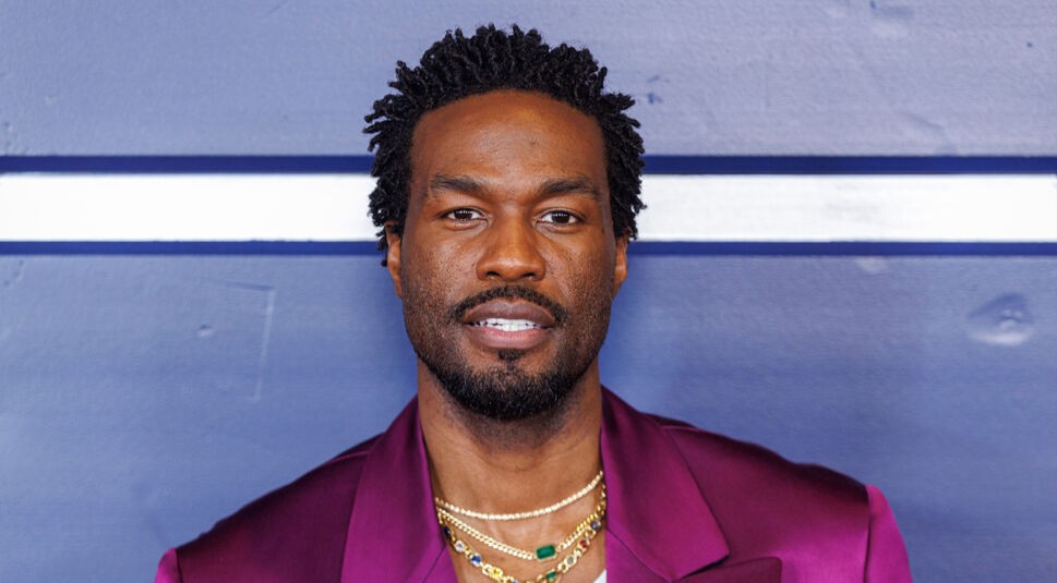 Auf dem Bild zu "Marvels Wonder Man" ist Yahya Abdul-Mateen II zu sehen. Er trägt ein auffälliges, purpurfarbenes Sakko über einem weißen Oberteil mit "Versace"-Schriftzug. Mehrere goldene Halsketten mit Edelsteinen vervollständigen seinen Look. Der Hintergrund ist schlicht blau mit einer weißen Linie, was den Fokus auf sein stilvolles Outfit und seine selbstbewusste Ausstrahlung lenkt.