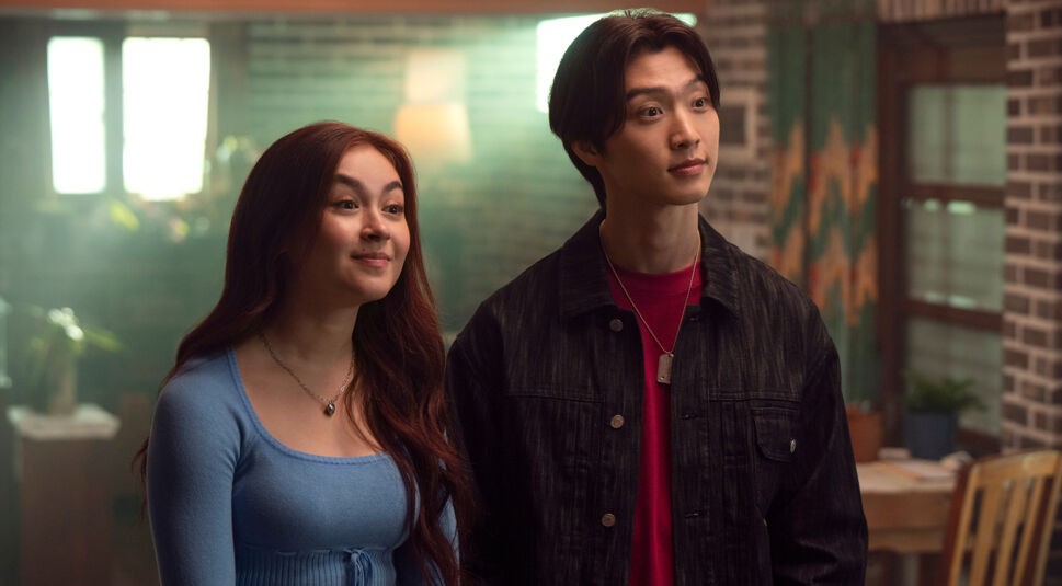 Auf dem Bild in dem Artikel zu XO, Kitty Staffel 3 stehen Kitty, gespielt von Anna Cathcart, und Min Ho, dargestellt von Sang Heon Lee, nebeneinander in einem gemütlich beleuchteten Raum. Kitty trägt ein himmelblaues Oberteil und lächelt leicht, während Min Ho in einer schwarzen Jacke mit rotem Shirt ernst, aber interessiert schaut. Die Szene wirkt warm und vertraut, mit Holzmöbeln und Pflanzen im Hintergrund.