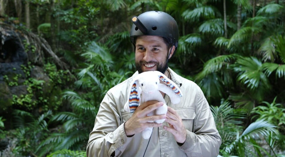 Auf dem Bild aus dem Dschungelcamp 2025 ist Maurice zu sehen, wie er inmitten des dichten Dschungels lächelt. Er trägt einen schwarzen Helm mit einer angebrachten Lampe und hält ein weißes Stofftier mit langen, bunt gemusterten Ohren in den Händen. Sein beigefarbenes Hemd passt zur tropischen Umgebung mit grünen Pflanzen und Palmen im Hintergrund.