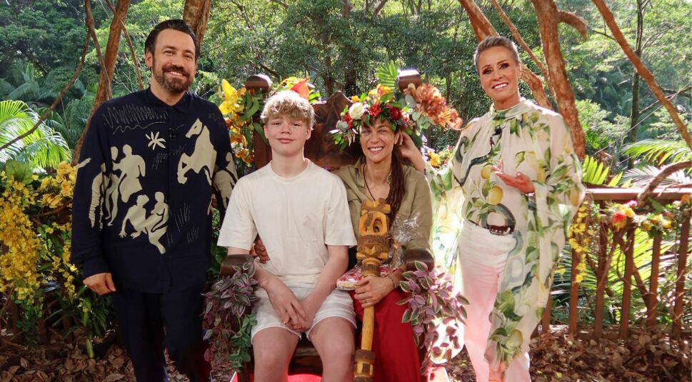 Auf dem Bild zum Dschungelcamp 2025 ist Lilly Becker als frisch gekrönte Dschungelkönigin zu sehen. Sie trägt eine bunte Blumenkrone und hält das Zepter in der Hand, während sie strahlend auf einem dekorierten Thron sitzt. Neben ihr sitzt ihr Sohn in einem weißen Outfit. Links und rechts von ihnen stehen die beiden Moderator:innen der Show, die ebenfalls lächeln. Die Szene ist von üppigem Dschungelgrün und farbenfrohen Blumen umgeben, die Stimmung ist feierlich und fröhlich.