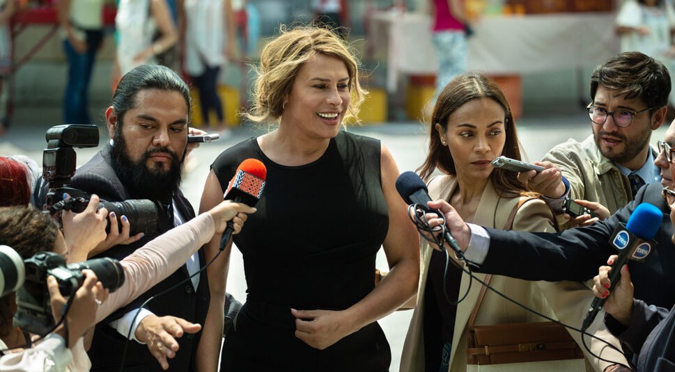 Auf dem Bild aus Emilia Pérez sind Karla Sofía Gascón (Mitte) und Zoë Saldaña (rechts) inmitten einer turbulenten Pressekonferenz zu sehen. Karla Sofía Gascón trägt ein elegantes schwarzes Kleid und lächelt selbstbewusst, während Mikrofone und Kameras auf sie gerichtet sind. Zoë Saldaña, in einem beigen Blazer, wirkt ernst und konzentriert. Die Szene vermittelt Spannung und öffentliche Aufmerksamkeit. Emilia Pérez streamen bietet die Möglichkeit, diesen Moment hautnah mitzuerleben.