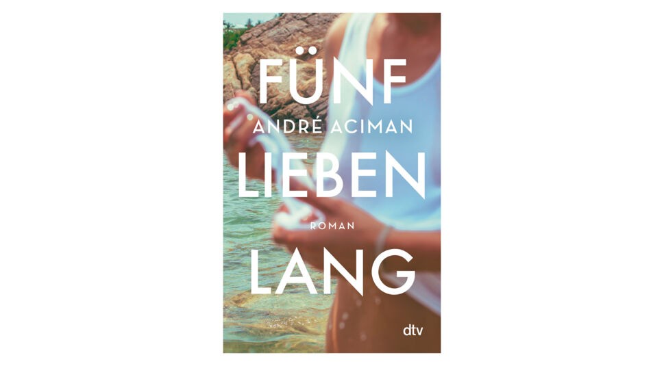 Auf dem Buchcover von Fünf Lieben lang von André Aciman, der Originalvorlage zur Serie Enigma Variations auf Netflix, ist eine verschwommene Szene am Wasser zu sehen. Eine Person in einem weißen Tanktop hält etwas in den Händen, während im Hintergrund Felsen und grünes Wasser zu erkennen sind. Die große, weiße Schrift dominiert das Cover und verleiht ihm eine sommerliche, nostalgische Stimmung.