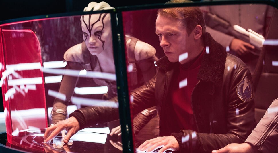 Auf dem Bild aus Star Trek Beyond, einem der Filme von Simon Pegg ist der Schauspieler in einer dramatischen Szene zu sehen. Er spielt Montgomery "Scotty" Scott und sitzt konzentriert an einer futuristischen Konsole mit holografischen Displays. Hinter ihm steht Jaylah, gespielt von Sofia Boutella, mit weißer Haut und schwarzen Streifen im Gesicht. Die Beleuchtung ist futuristisch mit roten und weißen Lichteffekten, was die angespannte Atmosphäre verstärkt.