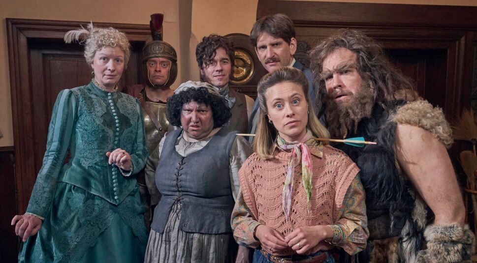 Auf dem Bild aus der deutschen Serie Ghosts stehen sieben Charaktere dicht beisammen in einem historischen Raum. Svenni (Sina Tkotsch) trägt moderne Kleidung und hat einen Pfeil quer durch den Hals. Neben ihr steht Urs (Jan van Weyde) mit zotteligem Fell und wildem Bart. Links sind Adelheid (Antje Widdra) in einem grünen Kleid, Claudius (Max Giermann) in römischer Rüstung und Griet (Meltem Kaptan) mit grimmigem Blick. Dahinter Joachim (Sebastian Schwarz) und Friedrich (Alexander Khuon).