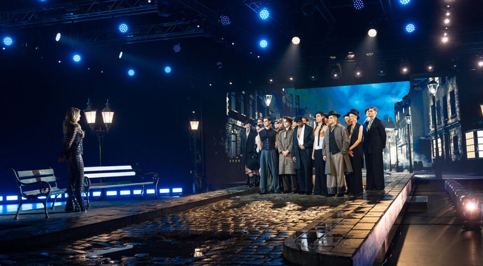 Auf dem Bild aus GNTM 2025 Folge 6 steht eine Frau in einem funkelnden schwarzen Outfit mit langen Ärmeln auf einer inszenierten Pflasterstraße, während sie einer Gruppe von Models gegenübersteht. Die Bühne ist im Stil einer nächtlichen Straßenszene mit Laternen, Bänken und einer großen Leinwand gestaltet, die eine historische Stadtlandschaft zeigt. Die Models tragen elegante Outfits mit Hüten, Westen und langen Mänteln. Die Beleuchtung sorgt für eine dramatische Atmosphäre.