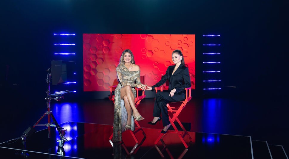 Auf dem Bild zur Show GNTM 2025 sitzen Heidi Klum und ihre Tochter Leni Klum auf roten Regiestühlen in einem modern beleuchteten Studio. Heidi trägt ein elegantes, schulterfreies Kleid mit Schlangenprint, während Leni einen schwarzen Hosenanzug mit hohen Schuhen trägt. Beide lächeln und halten sich an den Händen, während im Hintergrund ein rotes LED-Panel mit Wabenmuster leuchtet.