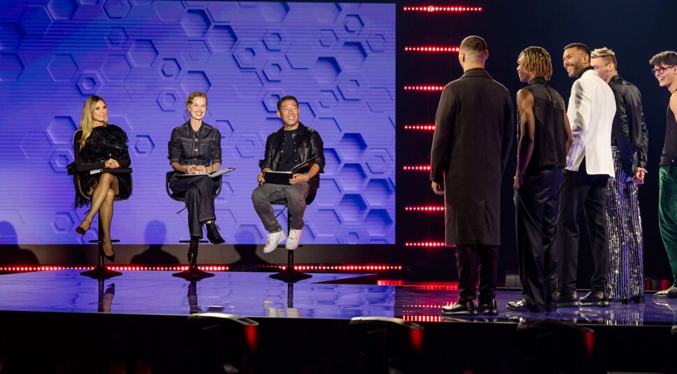 Auf dem Bild zu GNTM 2025 Folge 4 ist eine Jury mit drei Personen auf einer Bühne zu sehen, darunter Heidi Klum. Sie sitzen vor einem blauen Hintergrund. Ihnen gegenüber stehen mehrere männliche Models in unterschiedlichen Outfits. Die Bühne ist modern beleuchtet, mit roten LED-Streifen an der Seite. Die Stimmung wirkt professionell und gespannt.