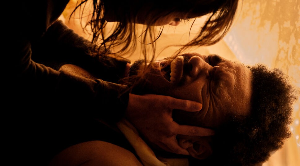 Auf dem Bild aus Hurry Up Tomorrow Film ist eine intensive Szene zwischen Jenna Ortega und Abel Tesfaye (The Weeknd) zu sehen. Jenna Ortega beugt sich mit nassen Haaren über Abel Tesfaye, hält sein Gesicht mit beiden Händen fest und blickt ihn eindringlich an. Tesfaye liegt auf dem Boden, sein Gesicht schmerzverzerrt, Schweißperlen auf der Stirn. Die warme, gedämpfte Beleuchtung verstärkt die emotionale und dramatische Spannung der Szene.