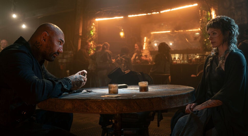 Das Bild in der In the Lost Lands Kritik zeigt eine stimmungsvolle Szene in einer düsteren Taverne, in der Boyce, gespielt von Dave Bautista, und Gray Alys, dargestellt von Milla Jovovich, an einem Tisch sitzen. 