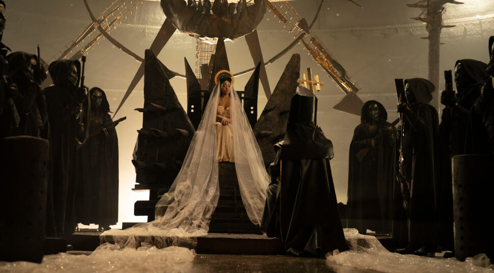 Das Bild in der In the Lost Lands Kritik zeigt eine eindrucksvolle Szene, in der die Königin Melange, gespielt von Amara Okereke, in einem prunkvollen goldenen Kleid mit langem Schleier auf einem dunklen Thron sitzt.