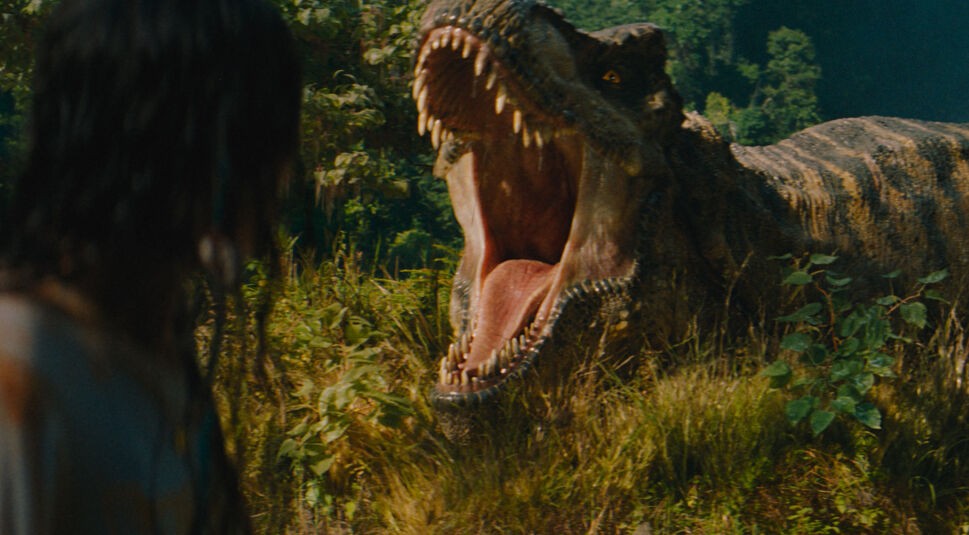 Auf dem Bild siehst Du eine Szene aus Jurassic World: Die Wiedergeburt. Luna Blaise steht im Vordergrund mit dem Rücken zur Kamera, während ein riesiger T-Rex im Hintergrund mit weit aufgerissenem Maul bedrohlich auf sie zustürmt.
