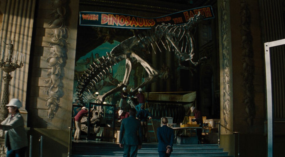 Auf dem Bild siehst Du eine Szene aus Jurassic World: Die Wiedergeburt. In der Empfangshalle eines Museums wird ein großes Dinosaurierskelett, vermutlich ein T-Rex, ausgestellt.