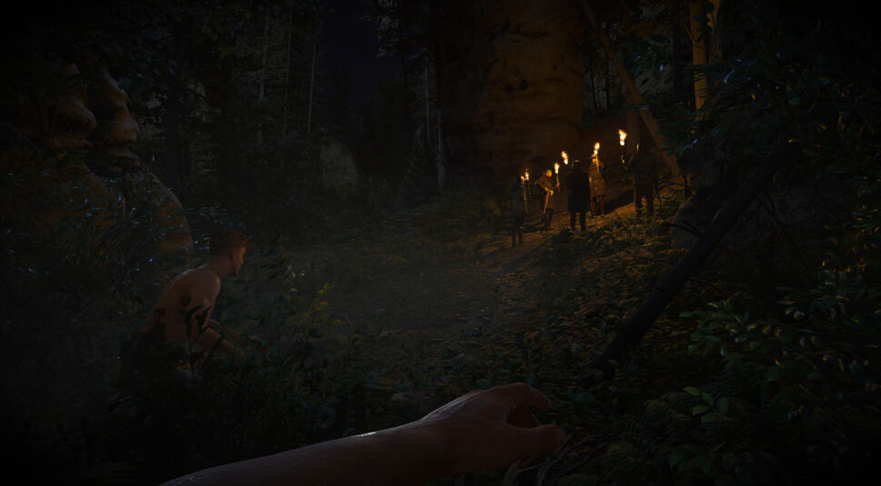 Auf dem Bild aus Kingdom Come Deliverance 2 ist eine nächtliche Schleichszene zu sehen. Der Spieler betrachtet aus der Ego-Perspektive eine Gruppe bewaffneter Männer mit Fackeln, die sich auf einem Waldpfad versammelt haben. Links kauert ein halbnackter Mann in den Büschen, offenbar versteckt. Das dunkle, dichte Waldgebiet mit geheimnisvollem Lichtspiel und tiefen Schatten erzeugt eine spannende, bedrohliche Atmosphäre.