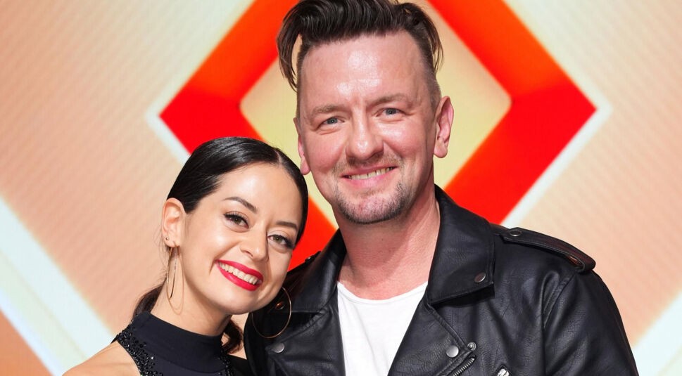 Auf dem Bild aus Let's Dance 2025 sind Ben Zucker und Malika Dzumaev in fröhlicher Stimmung zu sehen. Malika trägt ein elegantes schwarzes Kleid mit glitzernden Details und große goldene Creolen, während Ben eine schwarze Lederjacke über einem weißen Shirt trägt. Beide lächeln herzlich in die Kamera vor dem charakteristischen orangefarbenen Bühnenhintergrund der Show.