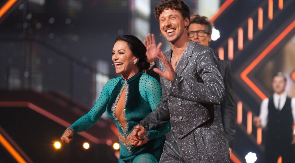 Auf dem Bild aus Let's Dance 2025 sind Christine Neubauer und Valentin Lusin in festlicher Stimmung auf der Bühne zu sehen. Christine trägt ein elegantes, smaragdgrünes Tanzkleid mit funkelnden Verzierungen und transparenten Ärmeln, während Valentin in einem silbernen Glitzeranzug strahlt. Beide lachen und wirken ausgelassen, während sie Hand in Hand jubeln. Im Hintergrund applaudiert das Publikum in einer stimmungsvollen, beleuchteten Kulisse.