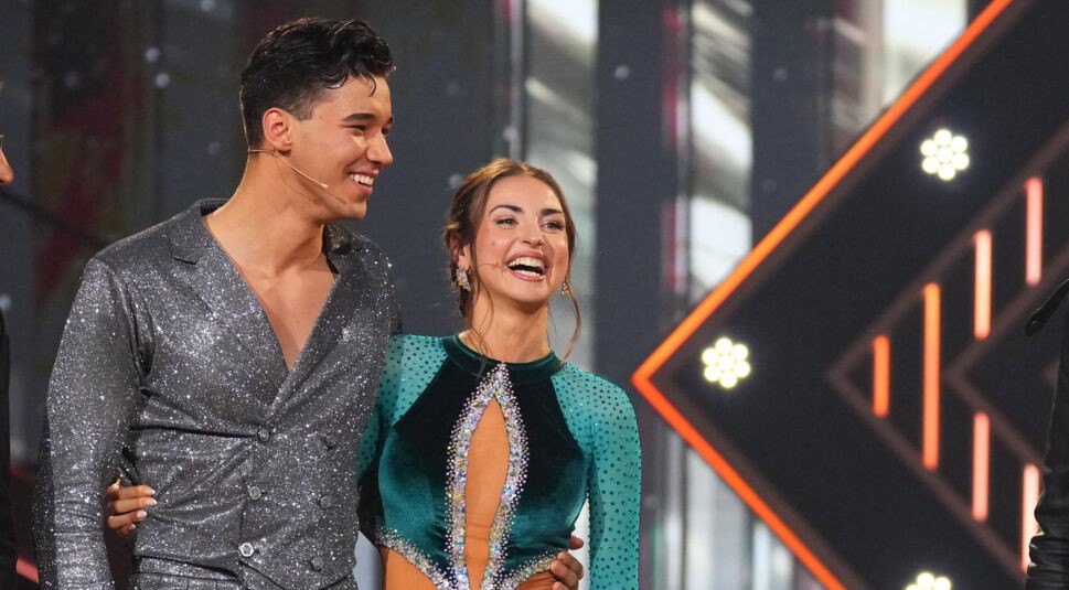 Auf dem Bild aus Let's Dance 2025 sind Diego Pooth und Ekaterina Leonova in strahlender Stimmung auf der Bühne zu sehen. Diego trägt einen funkelnden silbernen Anzug mit doppelreihigem Revers, während Ekaterina ein elegantes smaragdgrünes Tanzkleid mit glitzernden Verzierungen und transparenten Ärmeln trägt. Beide lachen glücklich, eng umschlungen, vor der atmosphärischen, beleuchteten Kulisse der Show.