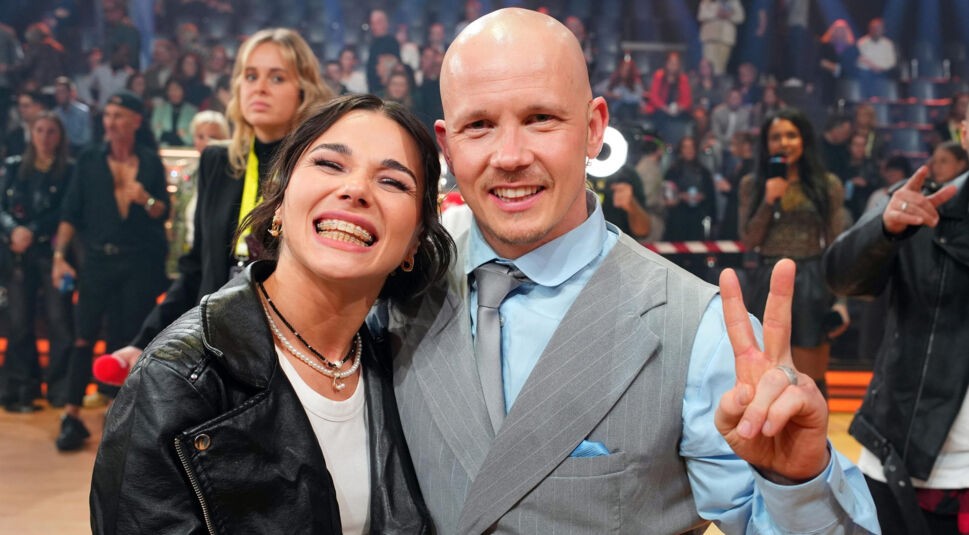 Auf dem Bild aus Let's Dance 2025 sind Fabian Hambüchen und Anastasia Maruster in ausgelassener Stimmung zu sehen. Fabian trägt einen eleganten grauen Anzug mit hellblauem Hemd und Krawatte und zeigt mit den Fingern das Victory-Zeichen. Anastasia trägt eine schwarze Lederjacke über einem weißen Shirt und lacht breit in die Kamera. Im Hintergrund ist eine lebendige Show-Atmosphäre mit Publikum und Moderatoren zu erkennen.