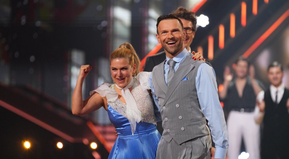 Auf dem Bild aus Let's Dance 2025 sind Jeanette Biedermann und Vadim Garbuzov in festlicher Tanzkleidung zu sehen. Jeanette trägt ein blaues, glitzerndes Kleid mit weißen, transparenten Ärmeln und zeigt eine jubelnde Geste mit erhobener Faust. Vadim steht neben ihr in einem eleganten grauen Anzug mit blauer Krawatte und lächelt strahlend. Im Hintergrund applaudiert das Publikum.