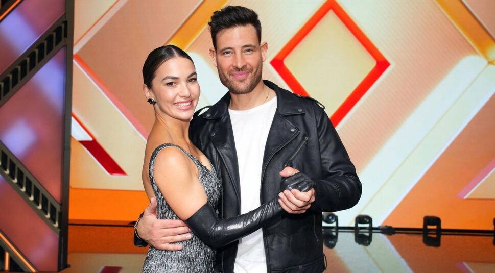 Auf dem Bild aus Let's Dance 2025 sind Marc Eggers und Renata Lusin in eleganter Tanzpose zu sehen. Renata trägt ein silbernes, glitzerndes Kleid mit schwarzen langen Handschuhen und lächelt strahlend. Marc kombiniert eine schwarze Lederjacke mit einem weißen Shirt und hält Renatas Hand in einer klassischen Tanzhaltung. Beide blicken glücklich in die Kamera vor dem stilvollen orangefarbenen Bühnenhintergrund.