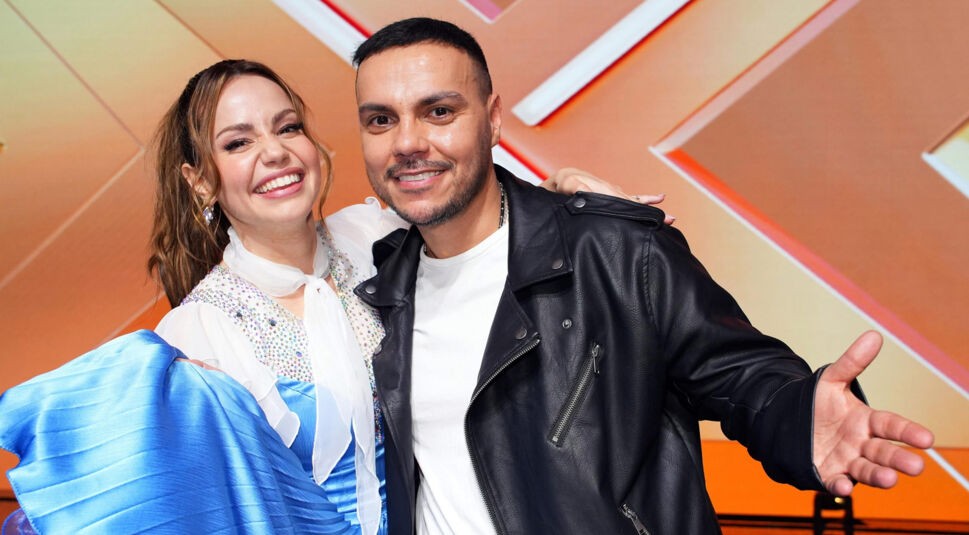 Auf dem Bild aus Let's Dance 2025 sind Osan Yaran und Christina Hänni in ausgelassener Stimmung auf der Bühne zu sehen. Christina trägt ein funkelndes blaues Tanzkleid mit weißen, transparenten Ärmeln und hebt fröhlich den Rock an. Osan ist leger in einer schwarzen Lederjacke über einem weißen Shirt gekleidet und breitet lachend die Arme aus. Beide strahlen in die Kamera vor dem orangefarbenen Bühnenhintergrund.