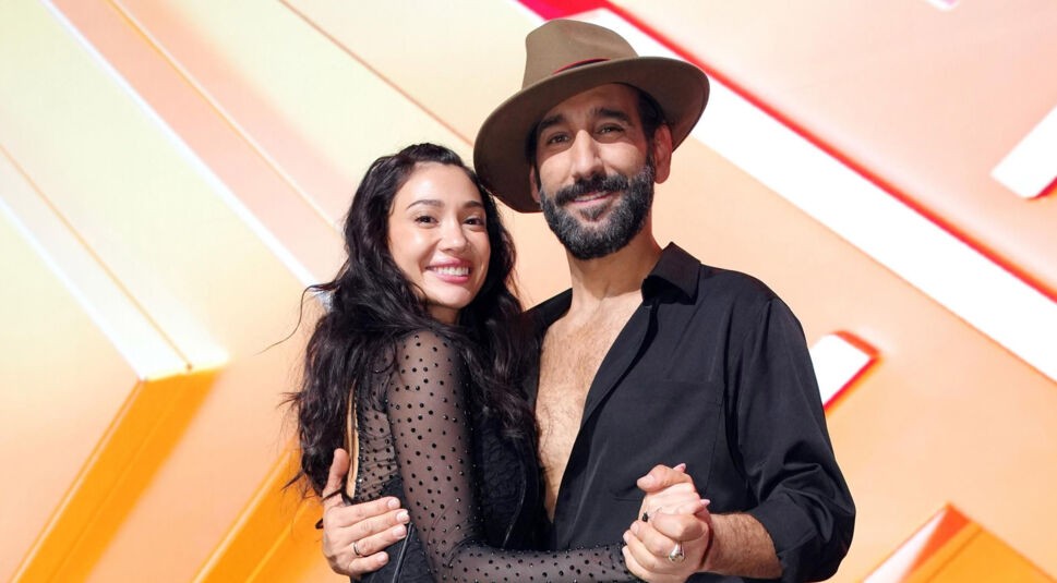 Auf dem Bild aus Let's Dance 2025 sind Paola Maria und Massimo Sinató in eleganter Tanzpose zu sehen. Paola trägt ein schwarzes, eng anliegendes Kleid mit transparenten, mit Strass besetzten Ärmeln und lächelt strahlend in die Kamera. Massimo trägt ein schwarzes, locker geöffnetes Hemd und einen braunen Hut, der seinen markanten Stil unterstreicht. Beide halten sich in einer Tanzhaltung vor dem orangefarbenen Bühnenhintergrund.