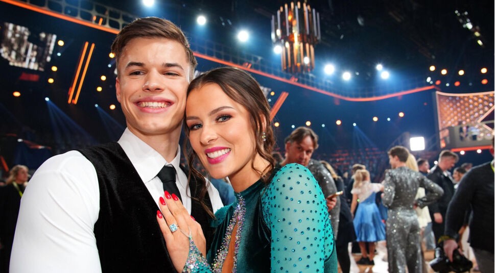 Auf dem Bild aus Let's Dance 2025 sind Taliso Engel und Patricija Ionel in festlicher Stimmung zu sehen. Taliso trägt ein klassisches Outfit mit weißem Hemd, schwarzer Weste und Krawatte, während Patricija ein elegantes smaragdgrünes Kleid mit glitzernden Verzierungen und transparenten Ärmeln trägt. Beide lächeln strahlend in die Kamera, während im Hintergrund die Tanzfläche mit weiteren Teilnehmern und der atmosphärischen Bühnenbeleuchtung zu sehen ist.