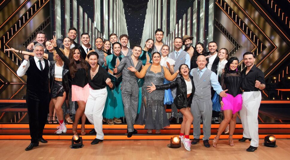 Auf dem Bild sind die Teilnehmenden von Let's Dance 2025 in festlicher Stimmung zu sehen. Die Gruppe besteht aus prominenten Tänzern und ihren Profi-Partnern, die in eleganten und farbenfrohen Outfits posieren. Die Bühne ist im Hintergrund mit Lichtelementen und modernen Designelementen gestaltet. Alle lächeln und zeigen ausgelassene Posen, was die energiegeladene Atmosphäre der Show widerspiegelt.