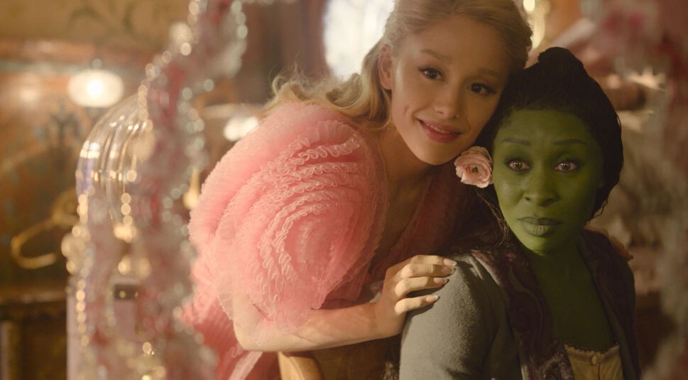 Auf dem Bild in dem Artikel zu den Oscar Filmen 2025, die bei GigaTV verfügbar sind, ist eine Szene aus Wicked: Teil 1 zu sehen. Zwei Frauen sitzen eng beieinander in einem warm beleuchteten Raum. Die Frau links trägt ein voluminöses, rosafarbenes Kleid und lächelt strahlend, während sie sich an die andere anlehnt. Die Frau rechts hat grüne Haut, dunkles Haar und einen leicht überraschten Ausdruck, mit einer rosa Blume im Haar. Die Szene wirkt magisch und freundlich.