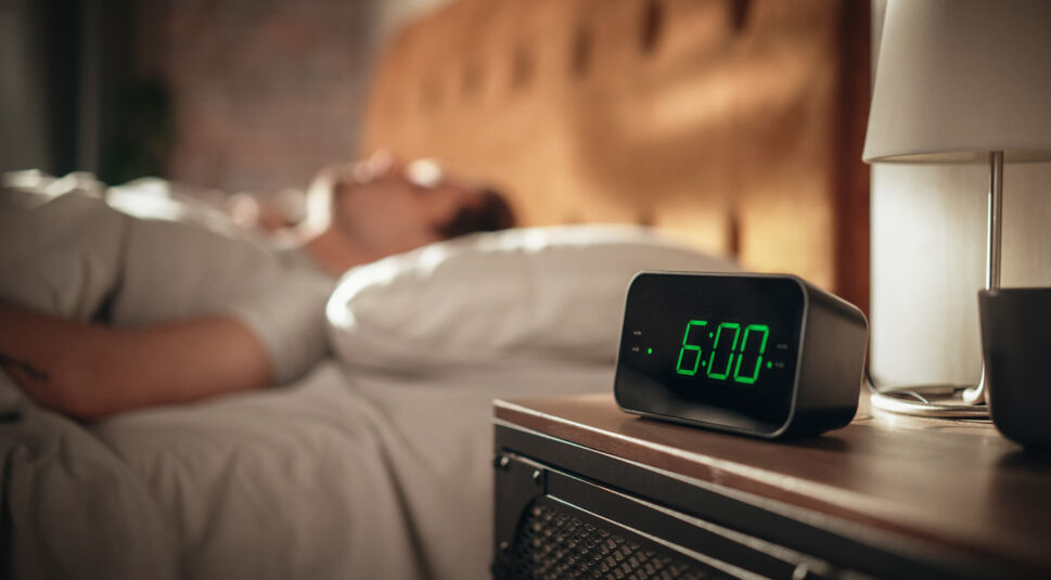 In einem gemütlichen Schlafzimmer zeigt ein digitaler Wecker auf dem Nachttisch 6:00 Uhr an. Im Hintergrund liegt ein Mann entspannt im Bett, während sanftes Morgenlicht den Raum erfüllt. Die Szene vermittelt Ruhe und Achtsamkeit und steht symbolisch für das Konzept "Phone Life Balance" – ein bewusster Start in den Tag ohne Ablenkung durch digitale Geräte.