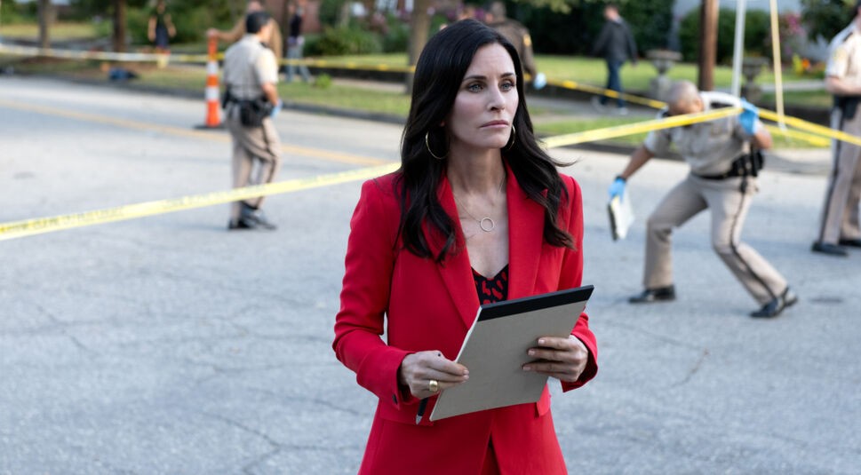 Auf dem Bild zu Scream 7 ist Courtney Cox als Gale Weathers zu sehen. Sie trägt einen auffälligen roten Blazer und hält ein Notizbuch in den Händen, während sie mit ernster Miene eine abgesperrte Tatortszene beobachtet. Im Hintergrund arbeiten Polizisten hinter gelbem Absperrband, was die dramatische und investigative Atmosphäre der Szene unterstreicht.