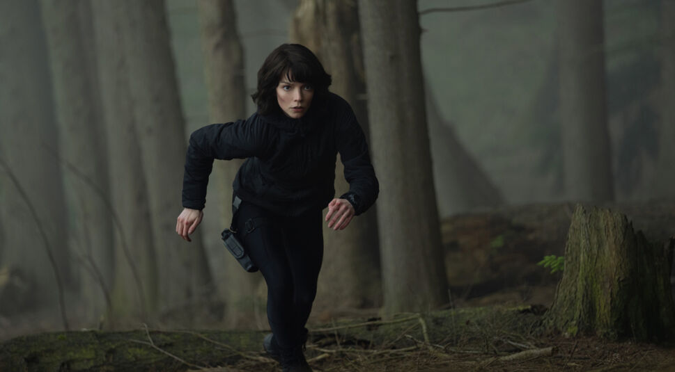 Auf dem Bild zu The Gorge Ende erklärt ist Anya Taylor-Joy als Drasa in einem nebligen Wald zu sehen. Sie trägt eine schwarze taktische Jacke und Hose, während sie mit konzentriertem Ausdruck nach vorne stürmt. Ihr dunkles Haar umrahmt ihr entschlossenes Gesicht, und eine Waffe ist an ihrem Oberschenkel befestigt. Die düstere, naturbelassene Umgebung mit moosbewachsenen Baumstümpfen und kahlen Ästen verstärkt die Spannung der Szene, die von Drasas Entschlossenheit und Dringlichkeit geprägt ist.