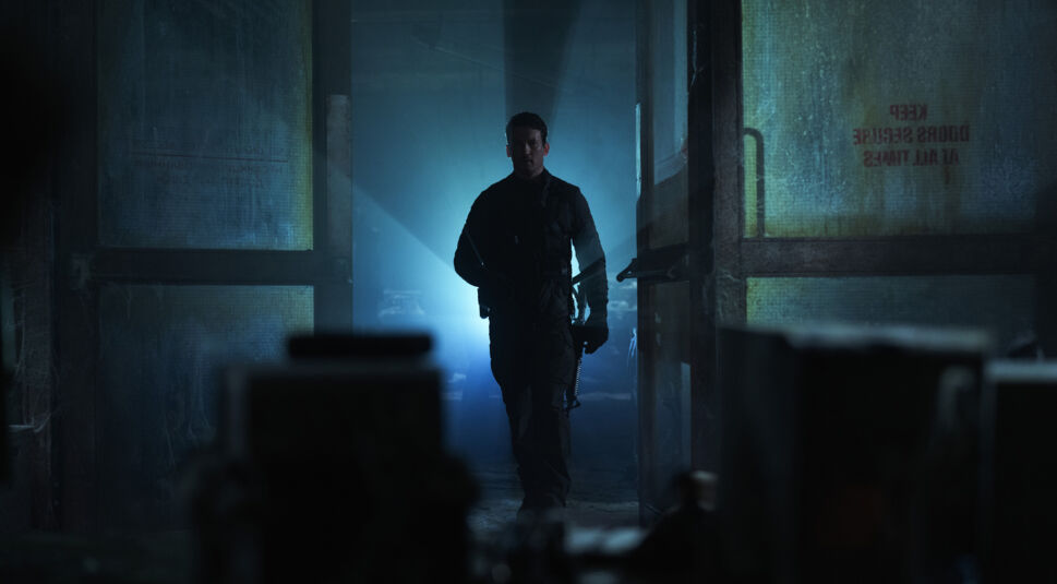 Auf dem Bild zu The Gorge Ende erklärt ist Miles Teller als Levi zu sehen. Er betritt einen düsteren, heruntergekommenen Raum durch eine offene Tür, während ein starkes blaues Licht von hinten seine Silhouette dramatisch hervorhebt. Er trägt eine taktische schwarze Ausrüstung und hält eine Waffe in der Hand. Die Umgebung wirkt verlassen und geheimnisvoll, mit staubigen Oberflächen und vergilbten Fenstern, auf denen eine Warnung aufgedruckt ist. Die Szene strahlt Spannung und eine bedrohliche Atmosphäre aus.