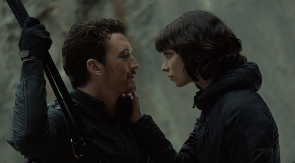 Auf dem Bild zu The Gorge Ende erklärt sind Anya Taylor-Joy als Drasa und Miles Teller als Levi in einem intensiven Moment zu sehen. Sie stehen sich nahe gegenüber, ihre Blicke tief ineinander versunken. Drasa hält Levis Gesicht sanft mit einer Hand, während er ein leichtes Lächeln zeigt. Levi trägt taktische Kleidung und hält ein Kletterseil, während Drasa eine schwarze Jacke trägt. Die Szene spielt sich in einer felsigen Umgebung ab und strahlt eine Mischung aus Spannung und emotionaler Verbundenheit aus.