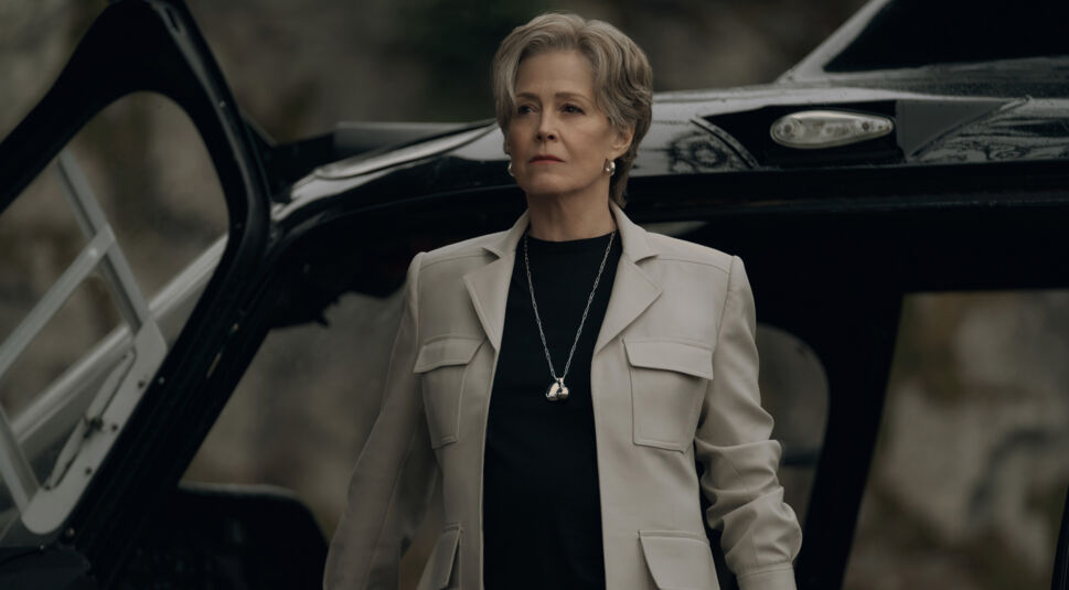 Auf dem Bild zu The Gorge Ende erklärt ist Sigourney Weaver als Bartholomew zu sehen. Sie steht entschlossen vor einem offenen Helikopter mit ernstem Gesichtsausdruck. Ihr silbernes Haar ist perfekt gestylt, sie trägt eine elegante cremefarbene Jacke über einem schwarzen Oberteil und eine lange Kette mit einem markanten Anhänger. Die düstere, verschwommene Naturkulisse im Hintergrund verstärkt die geheimnisvolle und autoritäre Ausstrahlung der Szene.