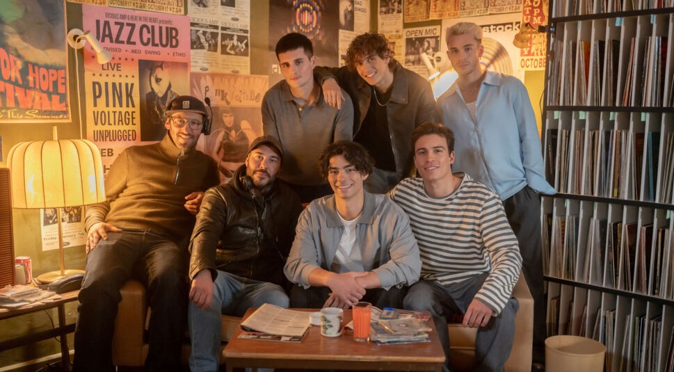 Das Bild zeigt die Darsteller von Back to the 90s beim entspannten Moment am Set. Sie sitzen und stehen in einem gemütlichen Raum mit Plattensammlung und Retro-Postern an der Wand. Julien, Tim, Luis, Jacob und Bene wirken gut gelaunt und locker, eingerahmt von Regisseuren und Crew. Die Szene strahlt kreative 90er-Jahre-Vibes aus.