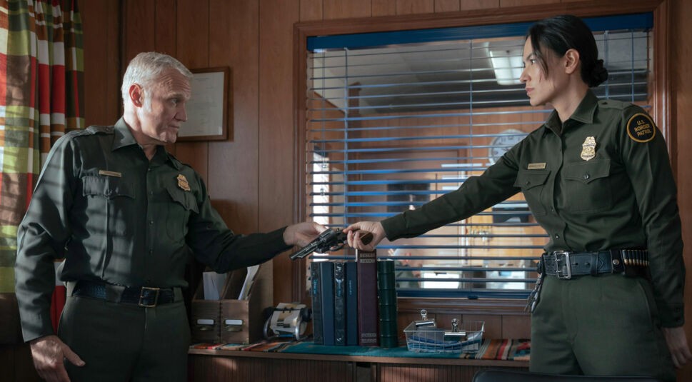 In Dark Winds Staffel 3 stehen Border Patrol Senior Chief Ed Henry (Terry Serpico) und Bernadette Manuelito (Jessica Matten) in einem Büro mit Holzvertäfelung. Beide tragen dunkelgrüne Uniformen der U.S. Border Patrol. Henry überreicht Manuelito mit ernstem Blick eine Pistole, während sie mit angespanntem Gesichtsausdruck danach greift.