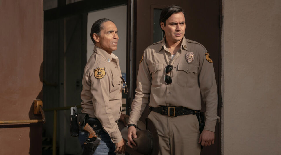 In Dark Winds Staffel 3 stehen Lt. Joe Leaphorn (Zahn McClarnon) und Jim Chee (Kiowa Gordon) in beigefarbenen Polizeiuniformen mit dem Emblem der Navajo Police vor einem Gebäude.